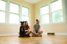 A man, woman, and dog at the end of the home-buying process, sitting on the wood floor of their new living space.