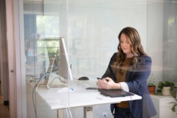 A professional woman at a standing desk, using her phone and computer to review her options trading plan.