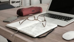 A desktop with a separately managed accounts ledger and laptop on it. On top of the ledger is a pair of reading glasses.