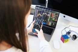 A woman sitting at a desk with her phone and laptop, conducting research before investing in stock