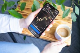 A hand holding a phone while the other hand holds a cup of coffee. The individual is reviewing their investment types on a finance app.