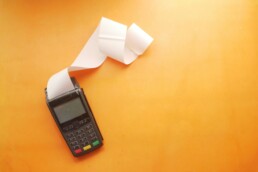 A calculating machine with long receipt paper coming out of it against an orange background. Used to calculate hidden costs in budgets